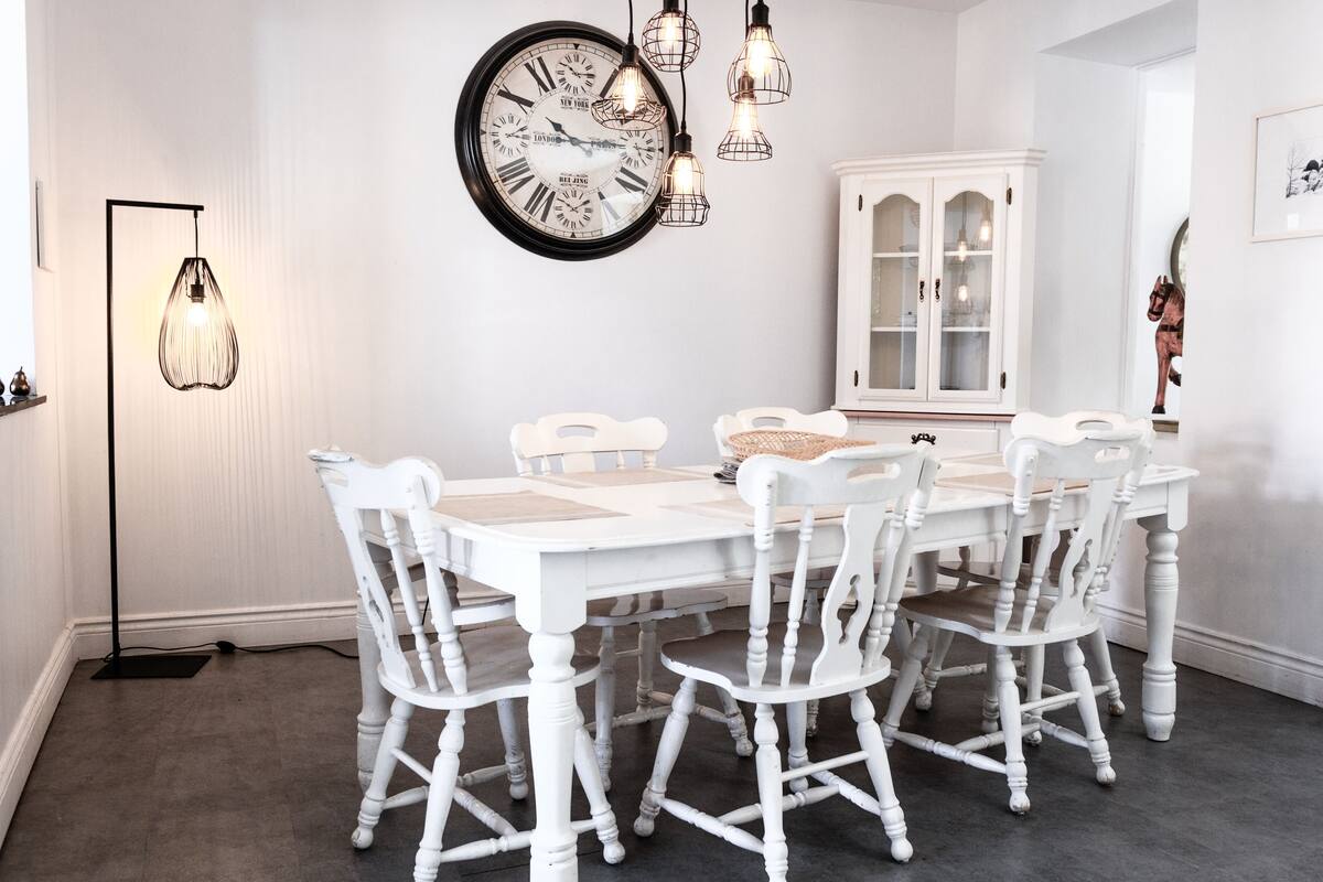 A dining room with a white table and chairs, a large wall clock, and pendant lights.