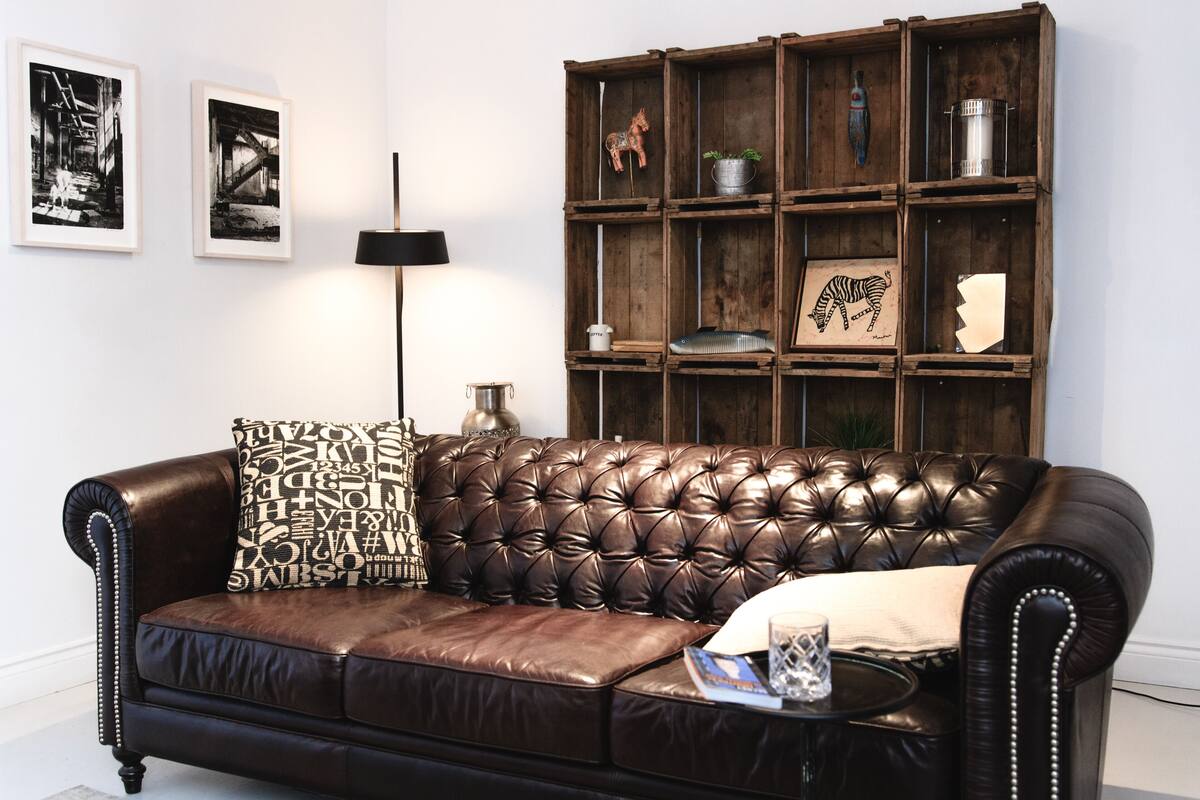 A brown leather sofa with a tufted backrest, a side table with a lamp, and a wooden shelving unit with various items.