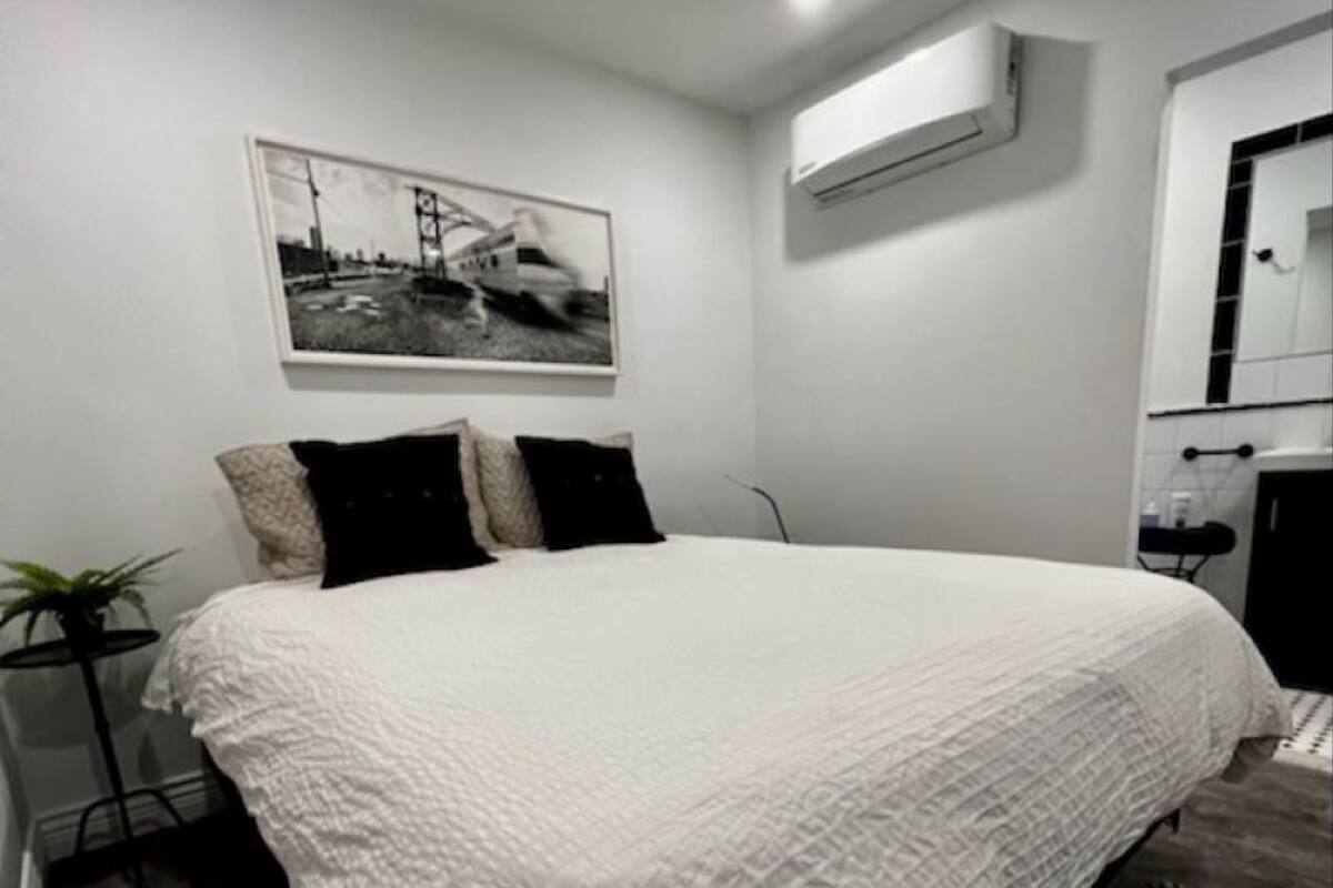A modern bedroom with a large bed, a small side table, a potted plant, and a framed picture on the wall.