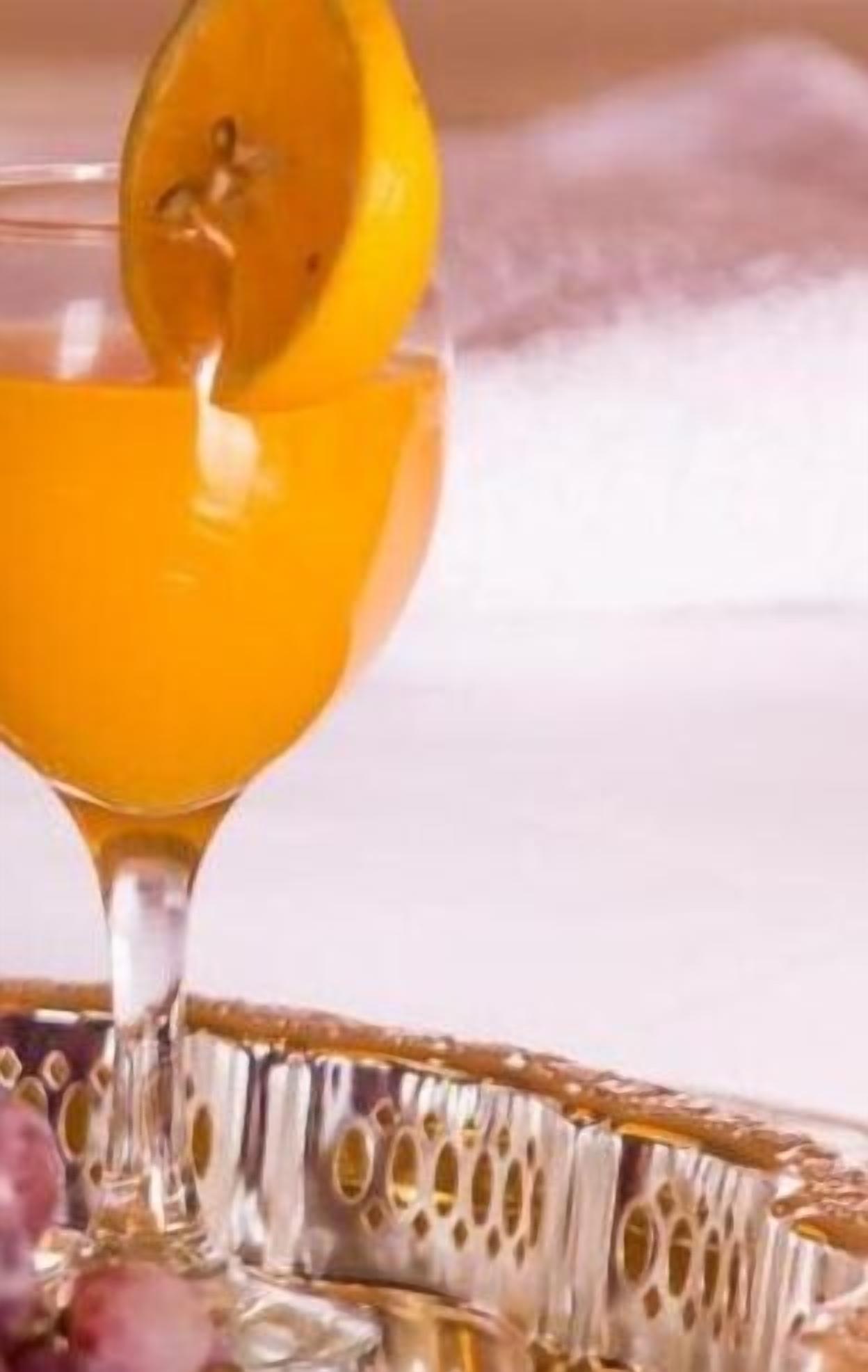 Un vaso transparente con borde dorado, lleno de un líquido naranja brillante, presumiblemente jugo, siendo servido desde una rodaja de naranja.