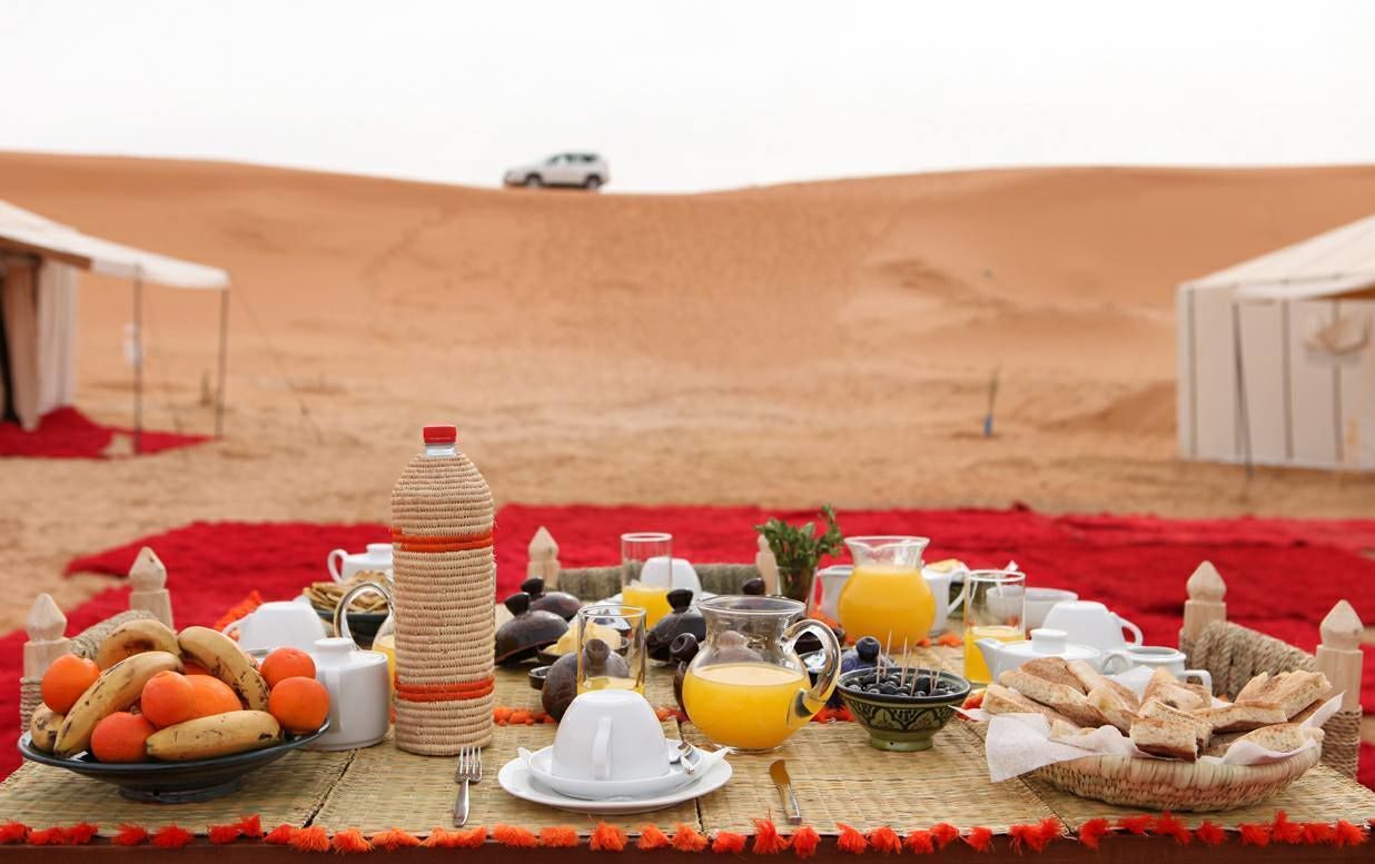A desert breakfast setup with food and drinks on a woven mat, tents in the background, and a vehicle on the sand.
