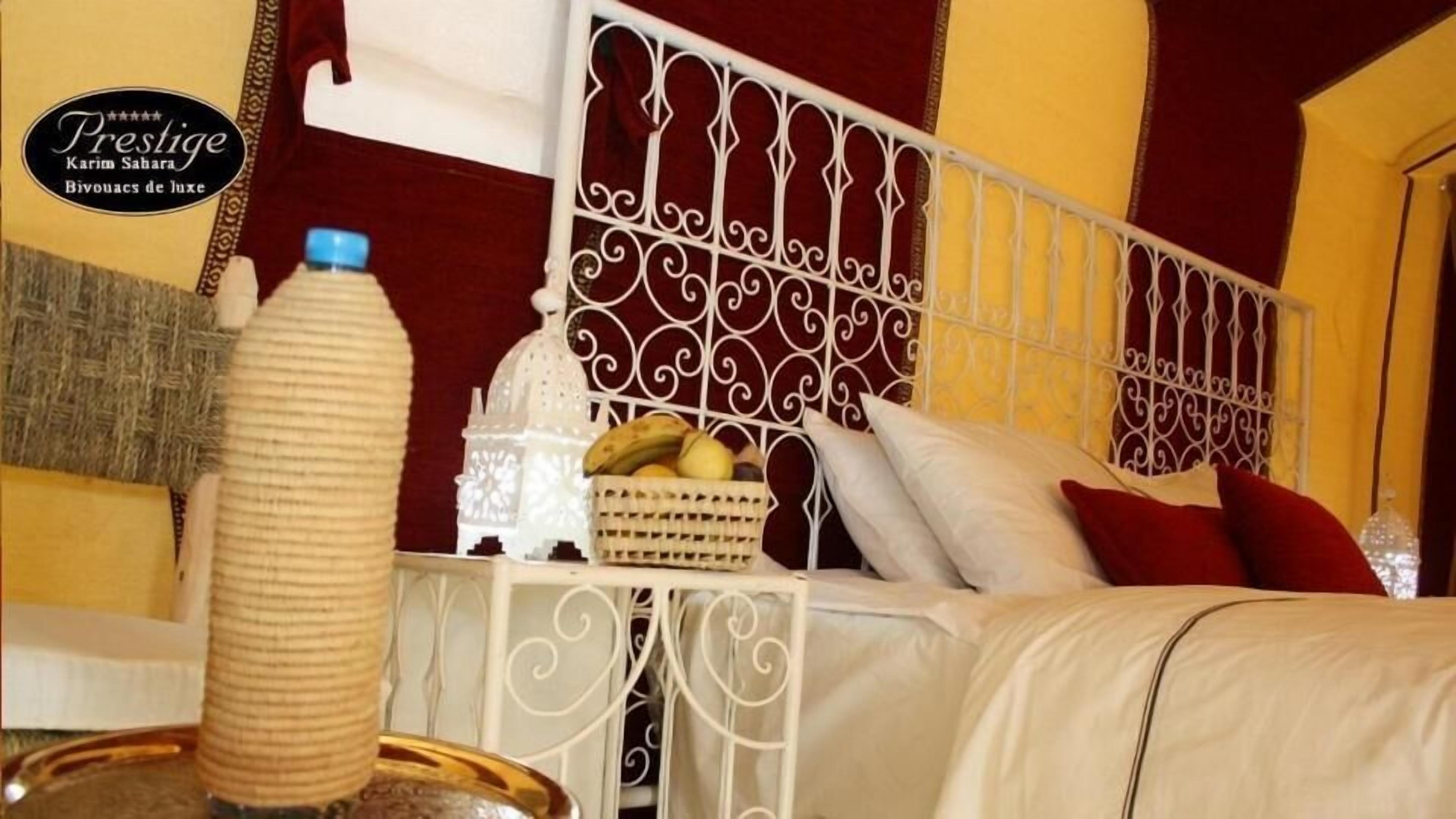 Ein Hotelzimmer mit einem dekorativen Metall-Kopfteil, einem Bett mit weißen und roten Kissen, einem Korbwesen mit Früchten und einer hohen, zylindrischen Vase.