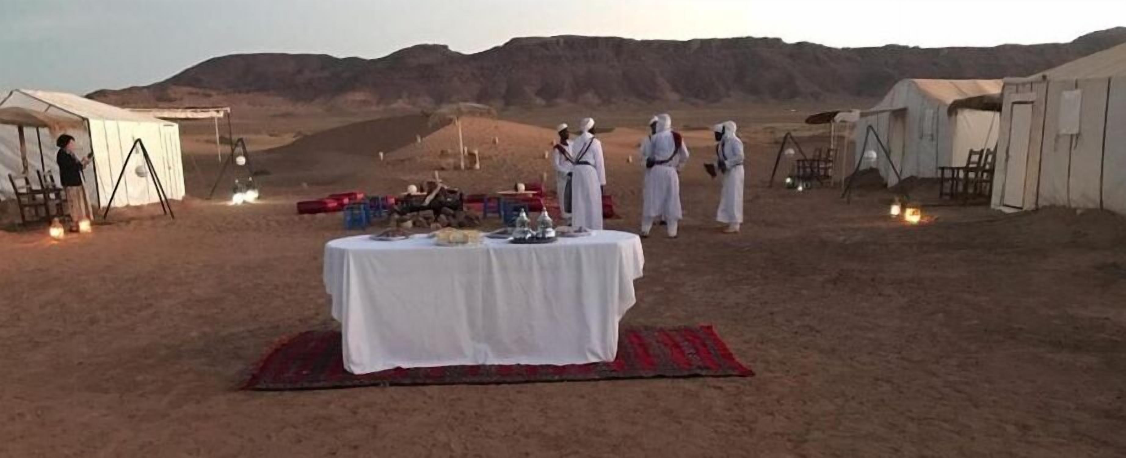 Campa en el desierto con tiendas de campaña, una mesa dispuesta para una comida y montañas al fondo.