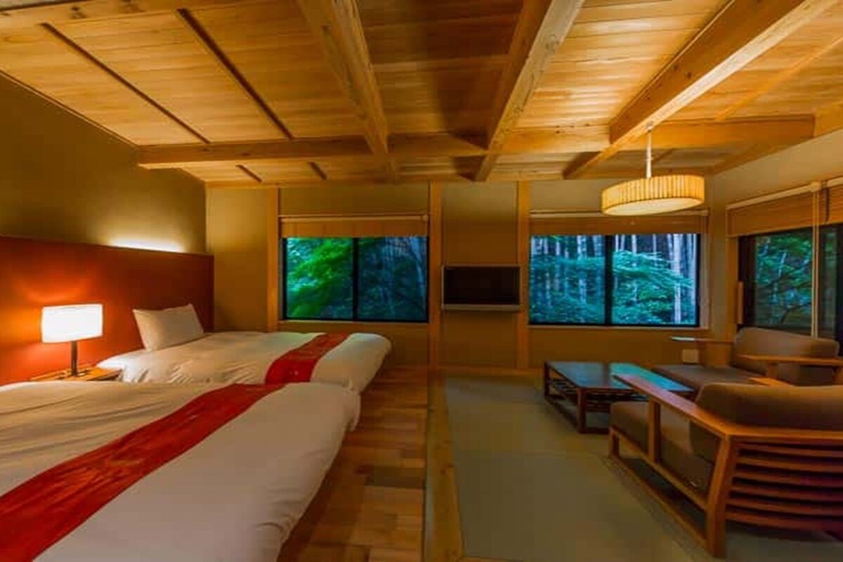 A hotel room with a large bed, a desk, a chair, and a TV. There are windows with a view of trees.