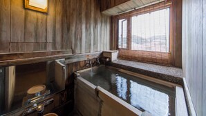 A rustic wooden bathroom with a large bathtub, a window with blinds, and a wooden shelf. - Tousin-an Komeya (Ito)