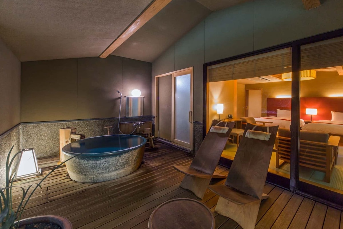 A hotel room with a large bed, a sitting area with a table and chairs, a bathtub, and a sliding glass door leading to a balcony.