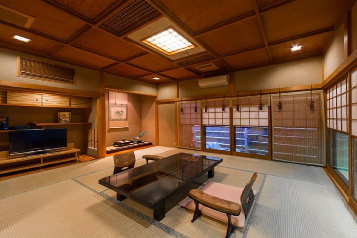 A traditional Japanese room with tatami flooring, a low table, a television on a wooden stand, and sliding doors with a view of the outside.