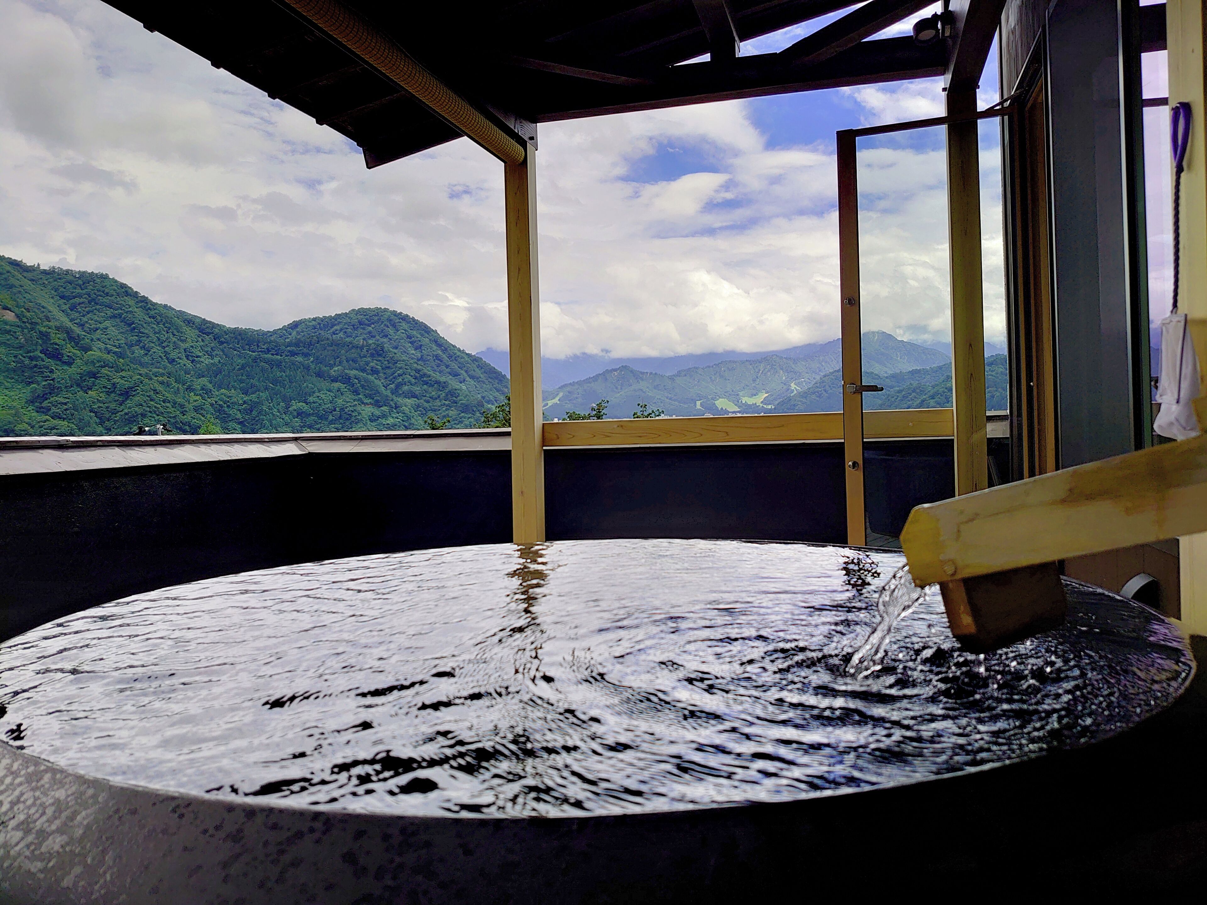 Una fuente termal con un banco de madera, vista a montañas y una puerta entreabierta.