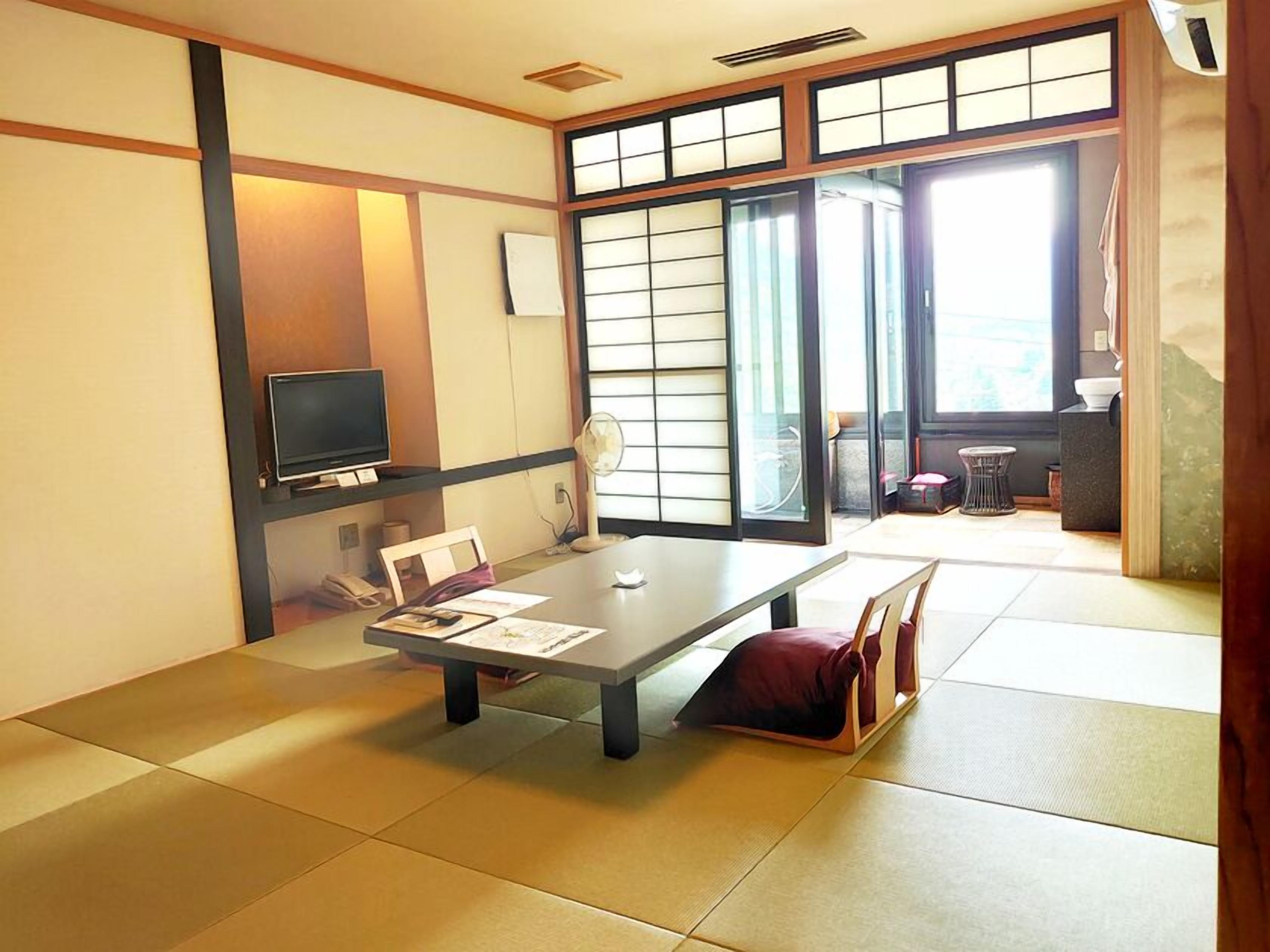 Ein traditioneller japanischer Raum mit Tatami-Boden, einem niedrigen Tisch mit Kissen, einem Fernseher und einer Schiebetür, die zu einem Balkon führt.