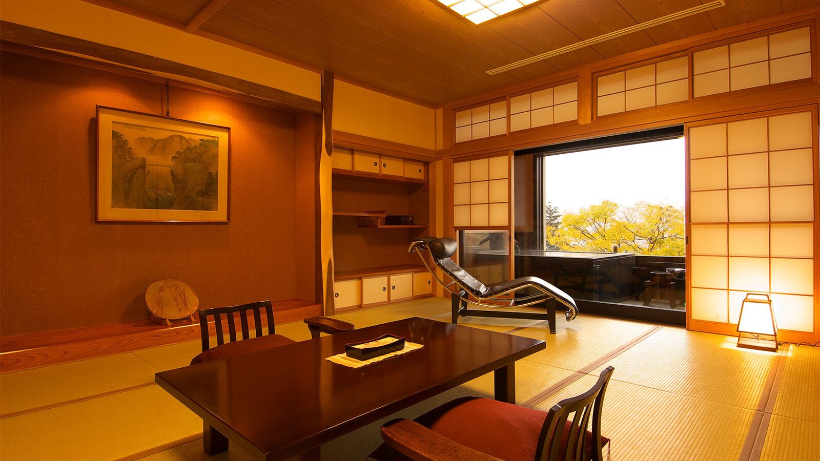 Ein traditioneller japanischer Raum mit Tatami-Bodenbelag, einem niedrigen Tisch, einem gepolsterten Stuhl, einer Schiebetür und einem gerahmten Bild an der Wand.