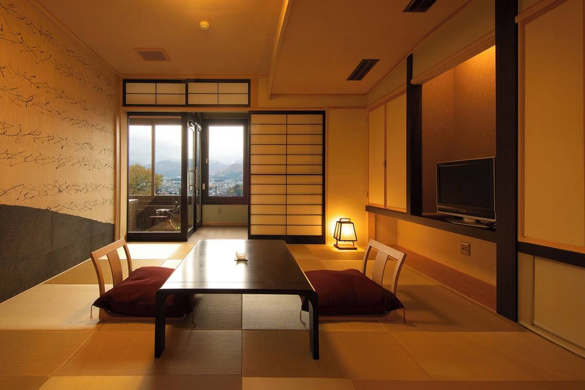 Una habitación japonesa tradicional con suelo de tatami, una mesa baja, puertas correderas, un televisor y vistas a un paisaje urbano.
