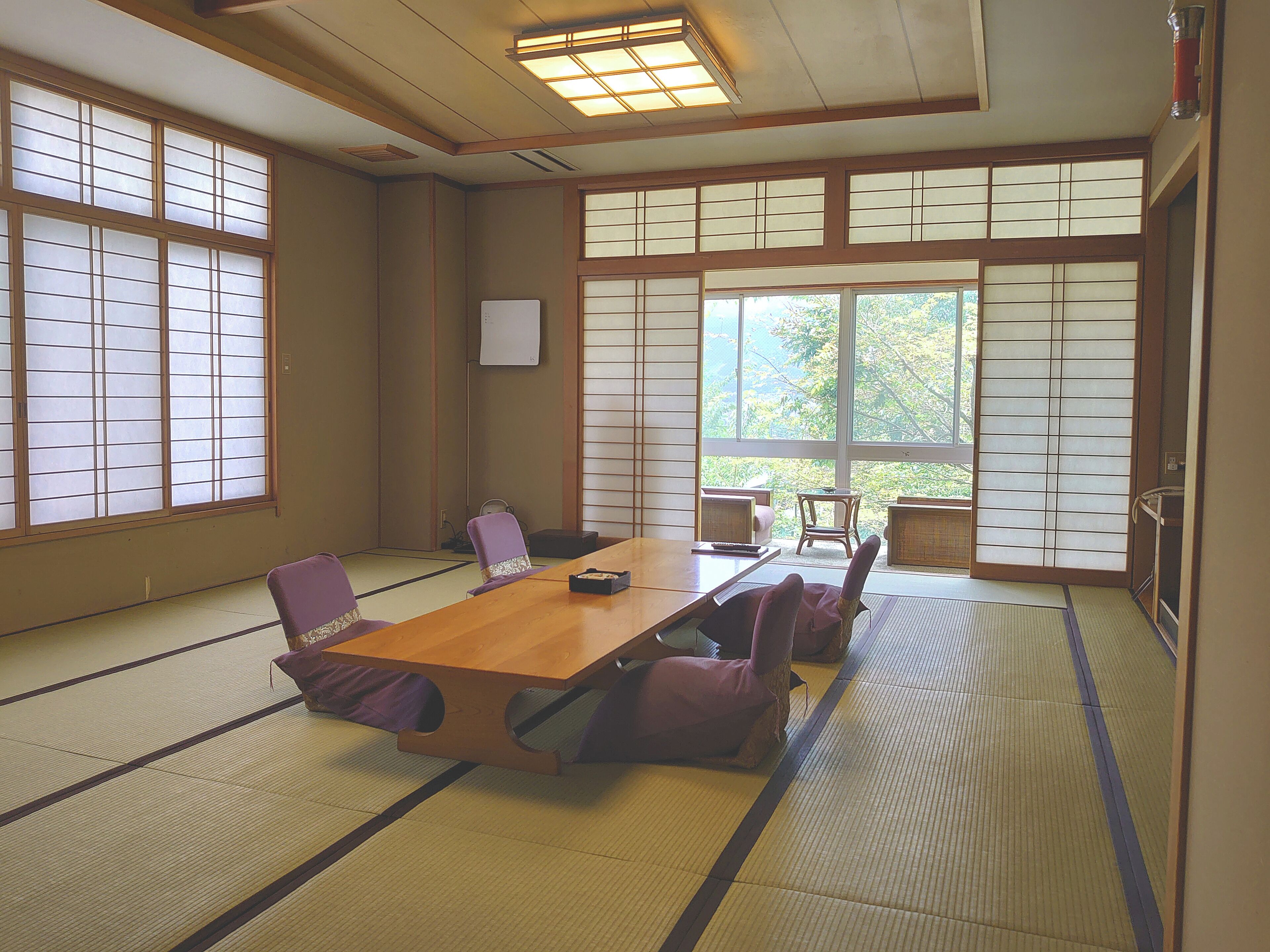 Ein Raum mit Tatami-Matten, einem Holztisch, violetten Kissen und großen Fenstern.