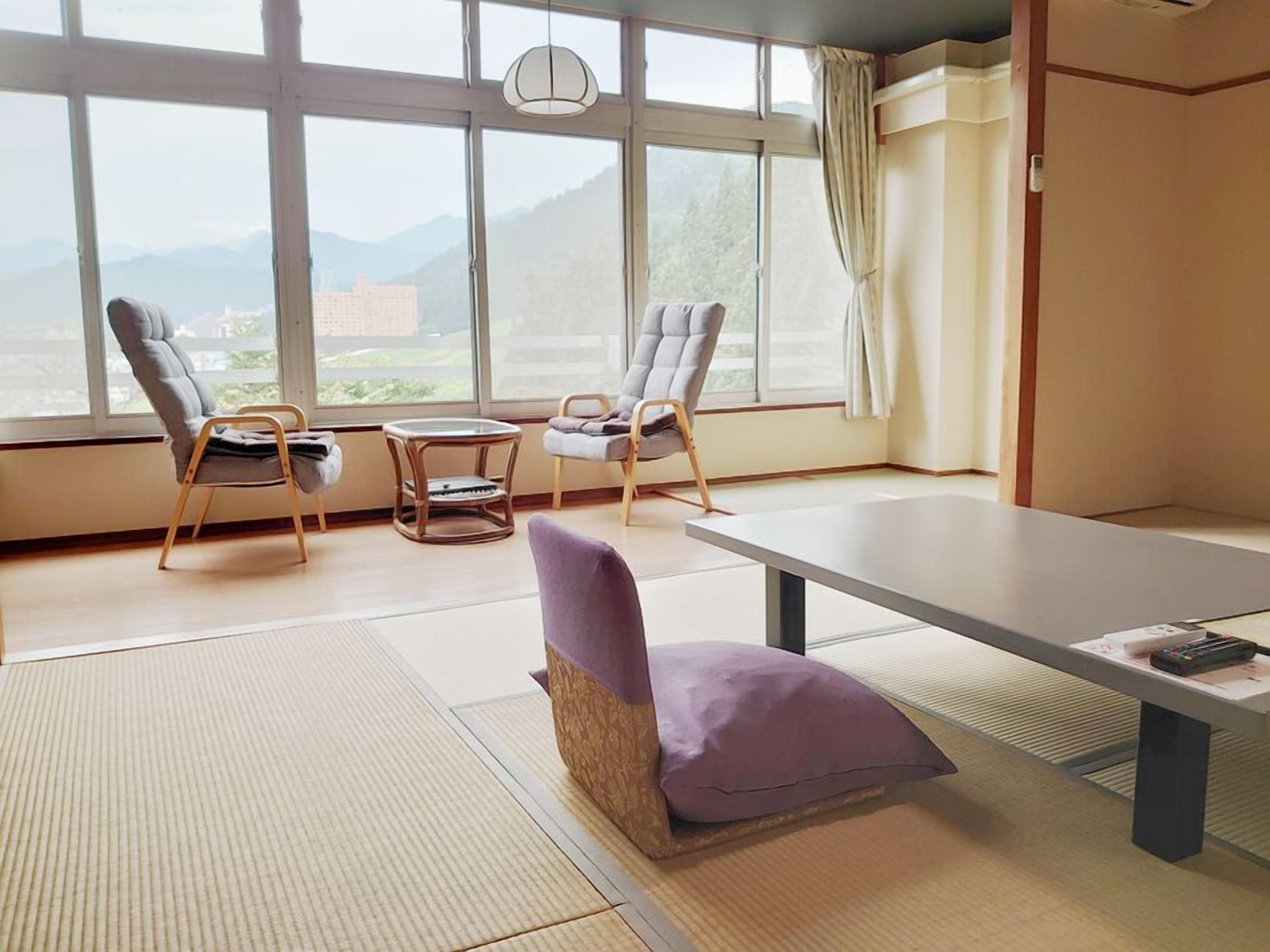 Ein Zimmer mit Tatami-Matten, einem niedrigen Tisch, zwei Stühlen und Blick auf Berge durch große Fenster.