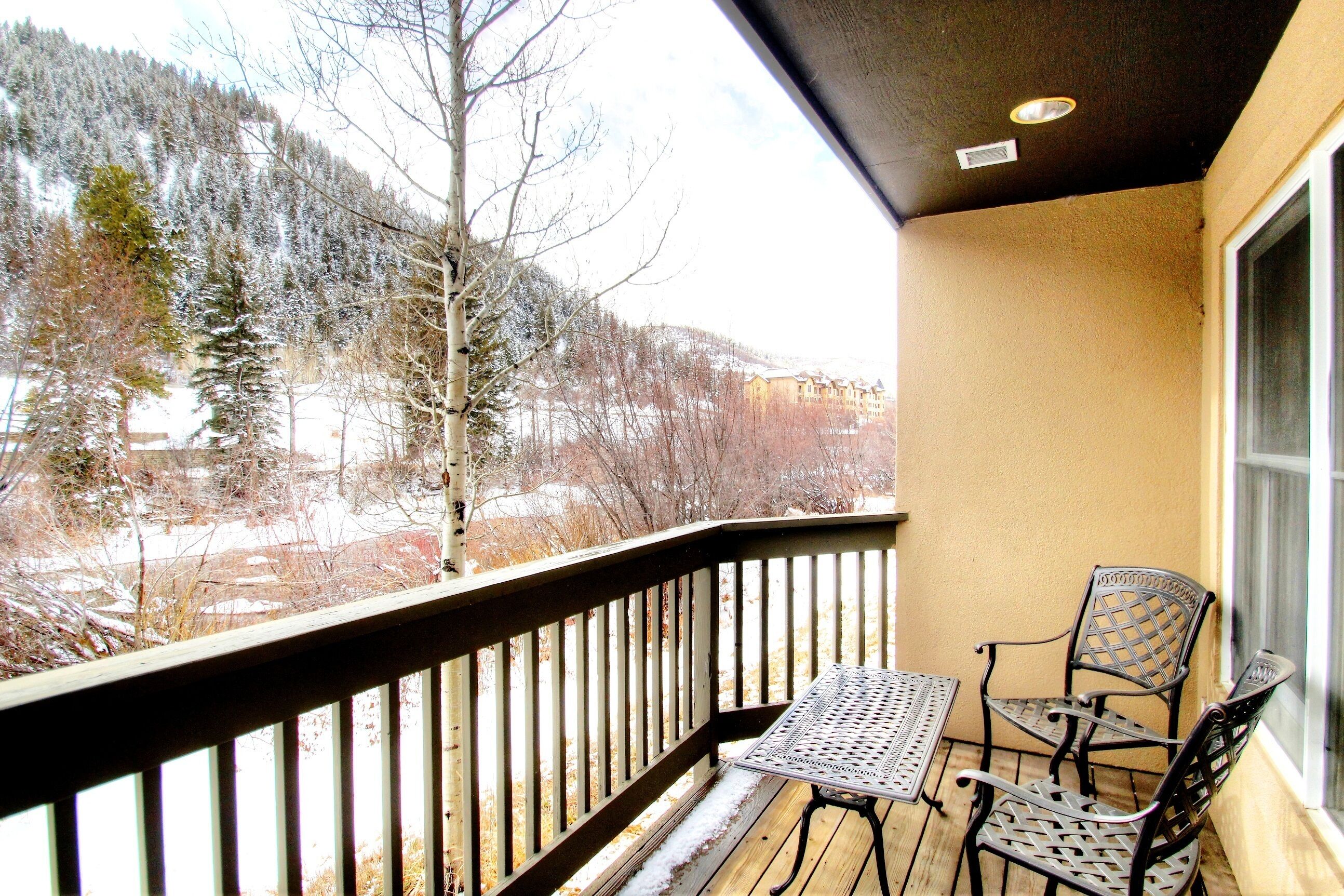 A balcony with a table and two chairs, overlooking a snowy landscape with trees and buildings.