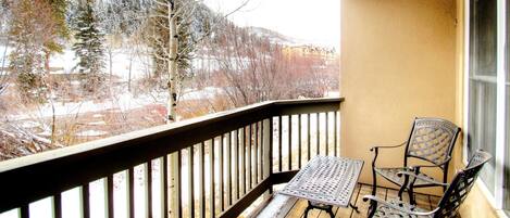 A balcony with a table and two chairs, overlooking a snowy landscape with trees and buildings.