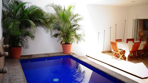 An outdoor swimming pool with two potted plants, a dining area with a table and chairs, and a white wall.