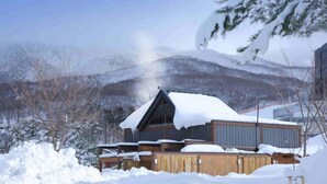 Eine Holzhütte mit geneigtem Dach, umgeben von schneebedecktem Boden und Bäumen, mit Bergen im Hintergrund.