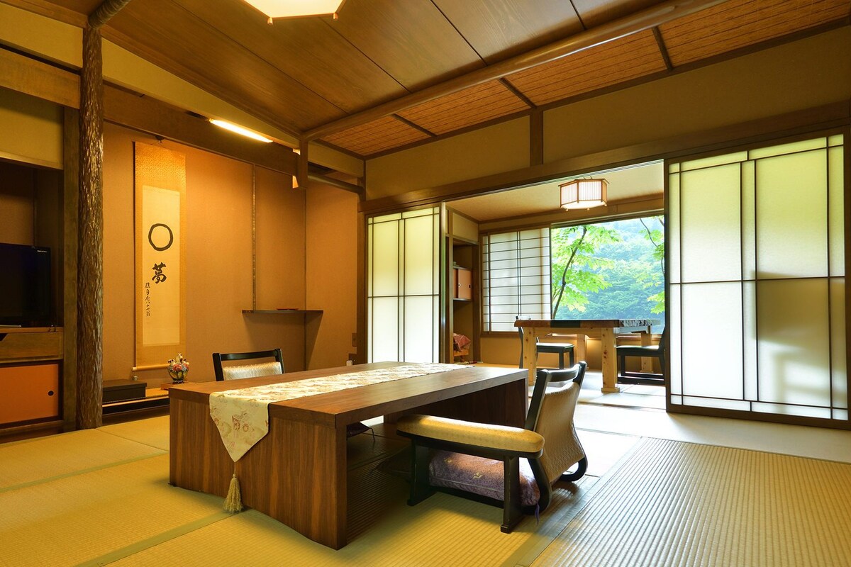 Une pièce de style japonais traditionnel, avec un sol recouvert de tatamis, une table basse, des chaises, un téléviseur, des portes coulissantes et une vue sur la verdure.