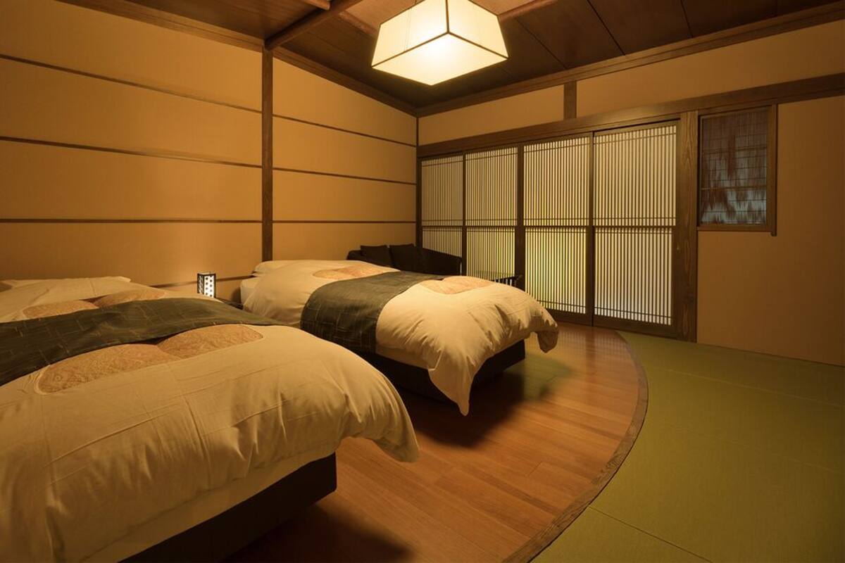 Una habitación tradicional japonesa con suelo de tatami, dos camas bajas con cojines, un divisor shoji y una lámpara colgante.