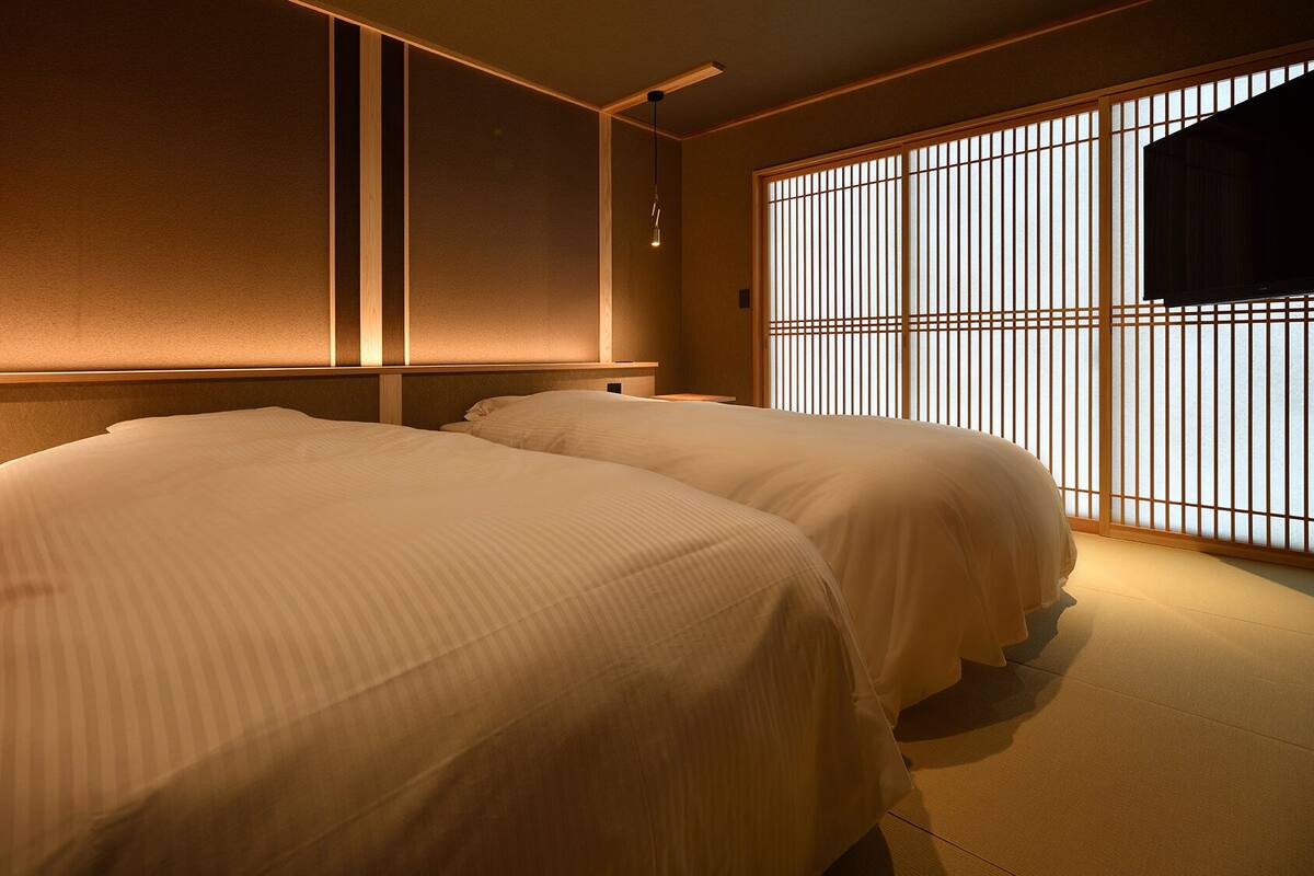 Habitación de hotel con dos camas, cabecera de madera, ventana grande con persianas verticales y televisor de pantalla plana.