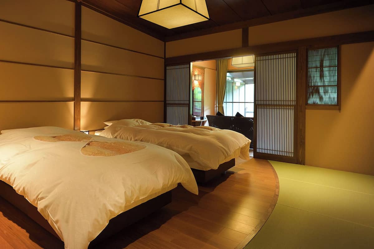 Un dormitorio de estilo japonés tradicional con suelo de tatami, dos camas con ropa de cama blanca, un divisor shoji y una lámpara de techo.