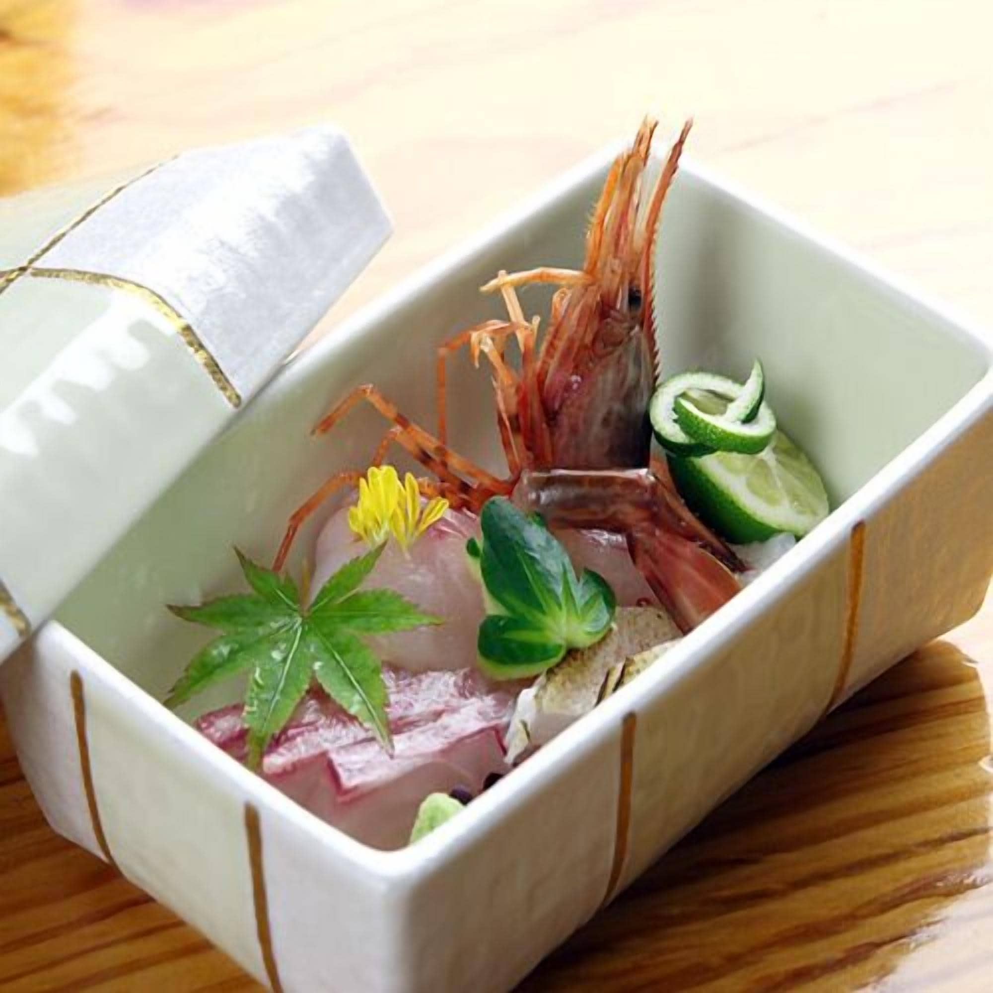 A rectangular container with a lid, containing a shrimp, slices of cucumber, a flower, and some green leaves.