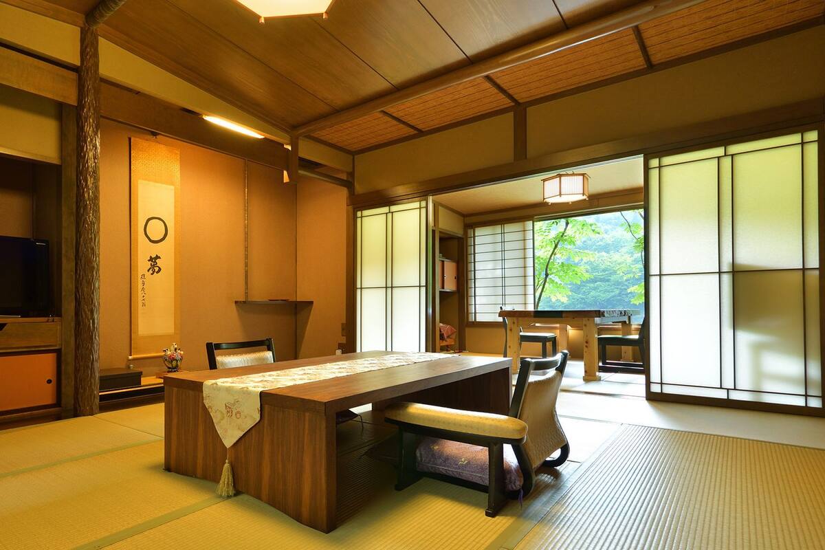 Una habitación tradicional japonesa con suelo de tatami, una mesa baja, sillas, un televisor, puertas correderas y vistas al verdor.