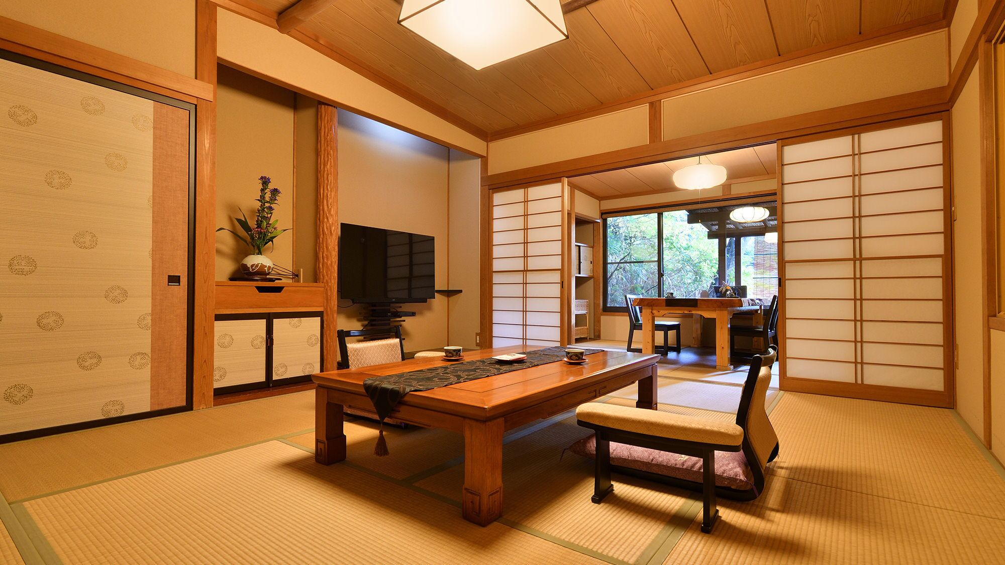 Une pièce de style japonais traditionnel, avec un sol recouvert de tatamis, une table basse, un téléviseur, des portes coulissantes et une vue dégagée sur l’extérieur grâce à une grande fenêtre.