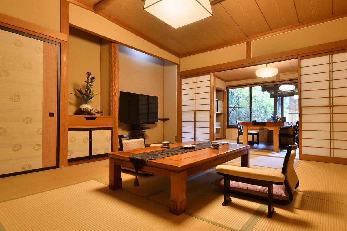 Una habitación japonesa tradicional con suelo de tatami, una mesa baja, un televisor, puertas correderas y vistas a través de un ventanal grande.