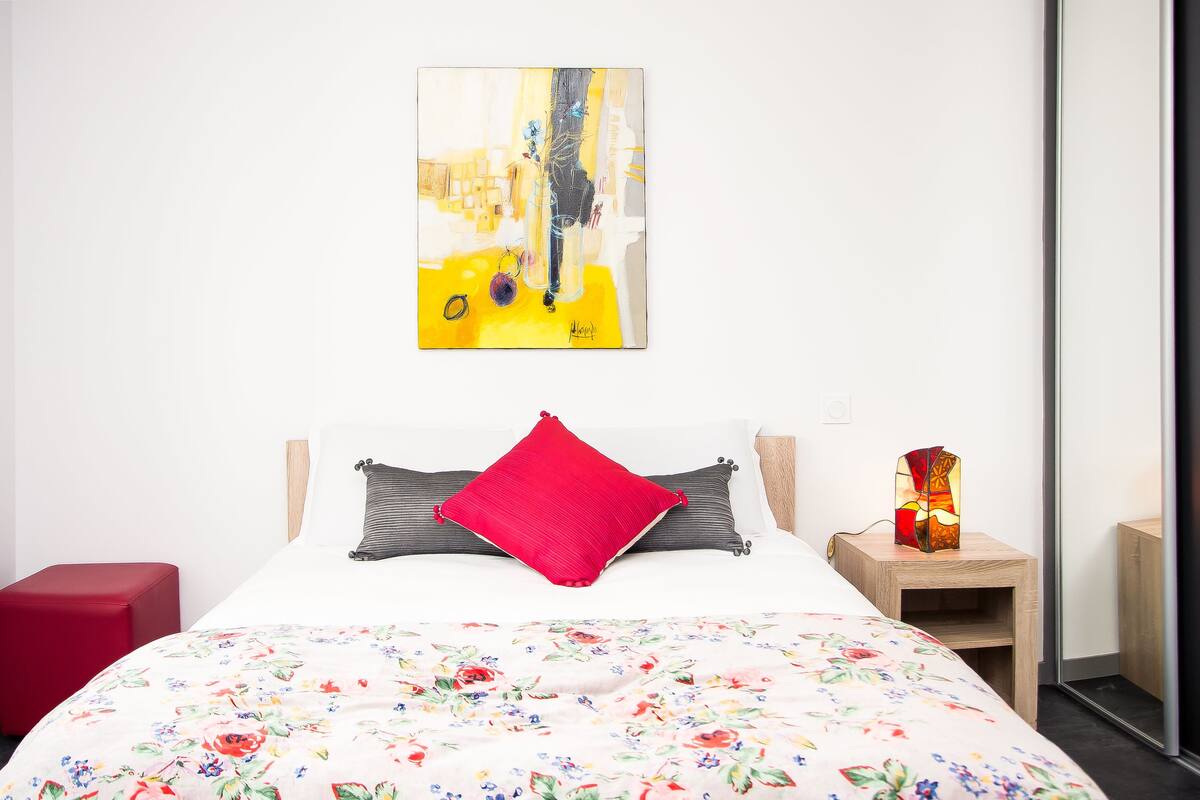 A bedroom with a floral bedspread, a red pillow, a painting on the wall, and a bedside table with a lamp.