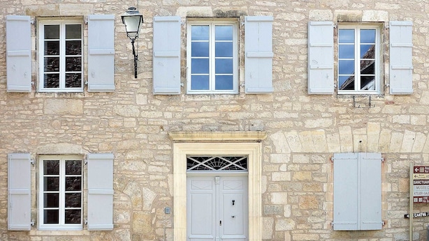 Un edificio de piedra con varias ventanas, contraventanas y una puerta central con una celosía metálica.
