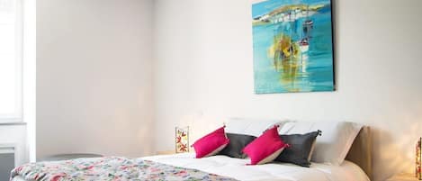 A neatly made bed with a floral bedspread and two pillows, a painting of a harbor scene on the wall, and a small bedside table with a lamp.