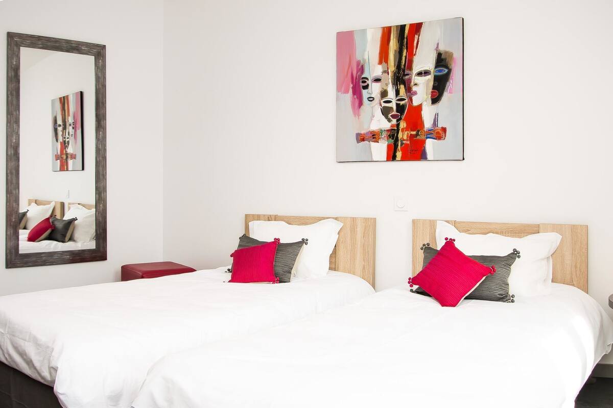 A hotel room with a large bed, two pillows, a red bedside table, and two wall-mounted paintings.