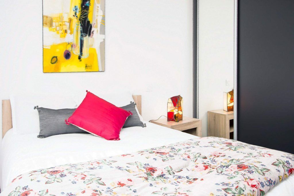 Une chambre à coucher avec un lit, un oreiller rouge, une couvre-lit à motifs floraux, une table de chevet avec une lampe et un tableau accroché au mur.