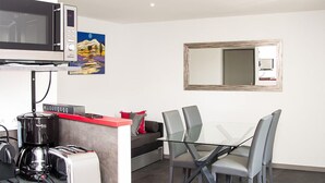 Una cocina moderna equipada con microondas, cafetera, tostadora y un refrigerador pequeño. Hay una zona de comedor con una mesa de vidrio y sillas.
