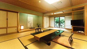 Quarto japonês tradicional com tatames, uma mesa baixa, cadeiras, uma TV e uma porta deslizante com vista para o verde.