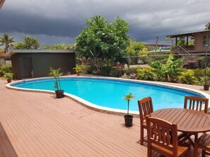 A swimming pool with a wooden deck, surrounded by potted plants and a wooden table with chairs.