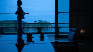 Silhouette einer Person stehend an einer spiegelnden Fläche, mit Blick auf ein Gewässer und Grünflächen, und Einblick in ein Gebäudefassade.