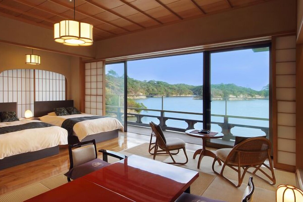 A hotel room with two beds, a dining table, chairs, a view of a lake, and a balcony.