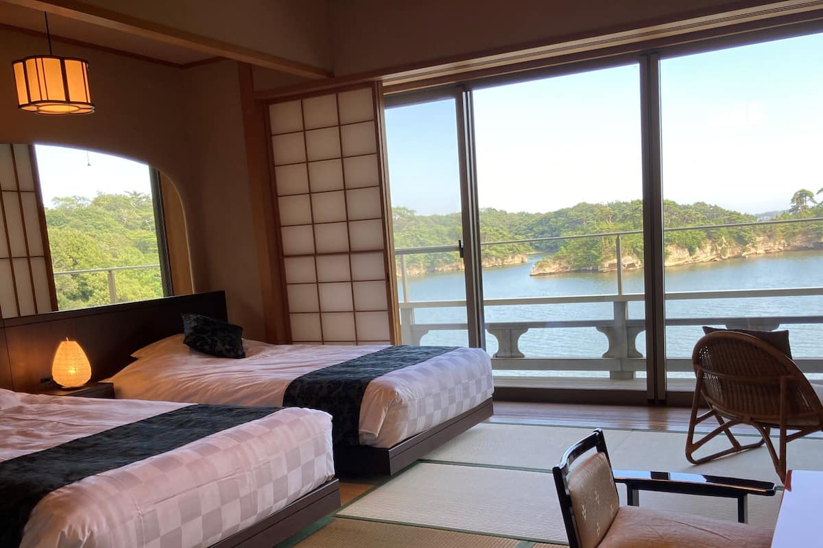 Habitación de hotel con dos camas, un ventanal con vistas a un lago, una silla y una mesa.