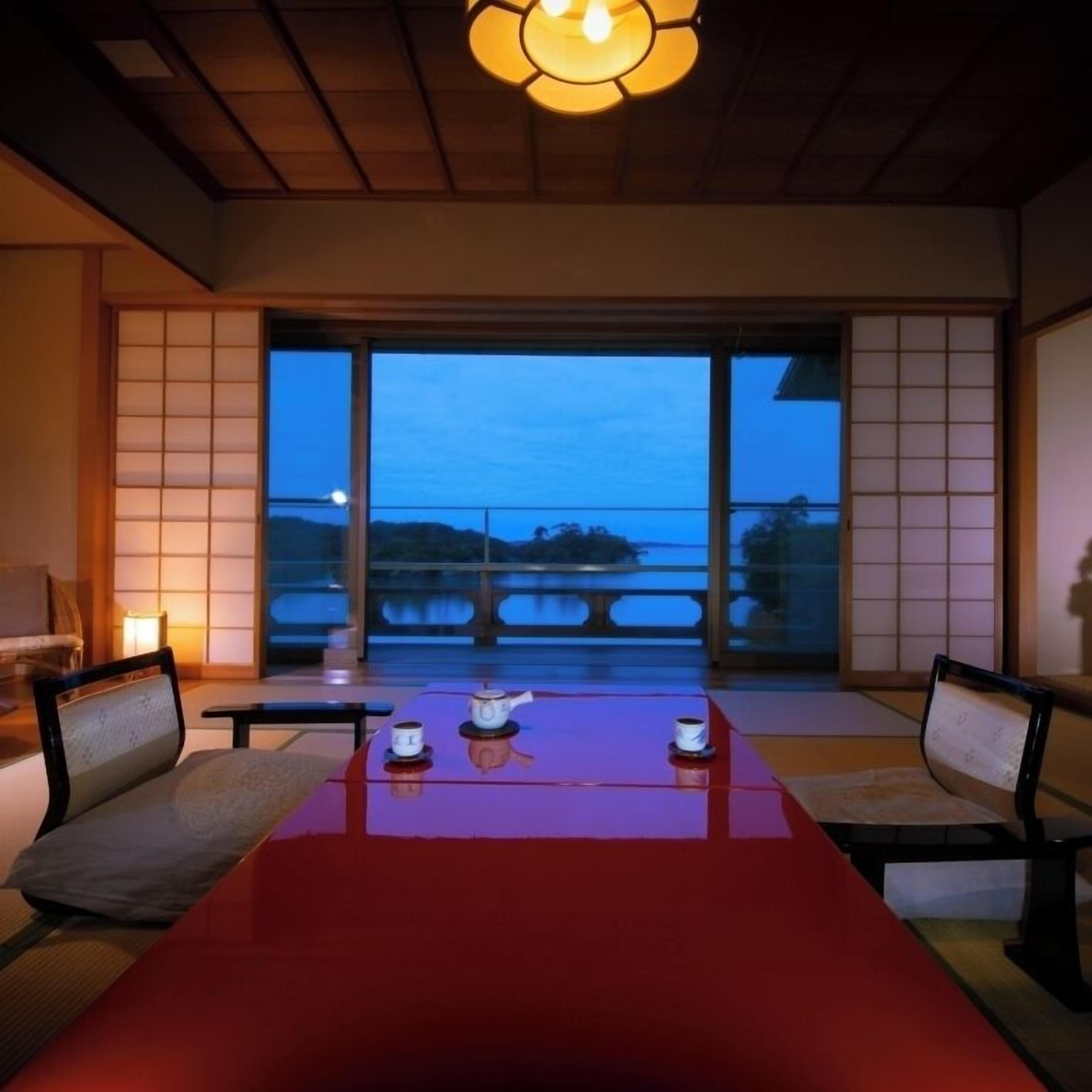 Una habitación con un gran ventanal corredero, una mesa roja con dos tazas y una tetera, y puertas correderas tradicionales japonesas.