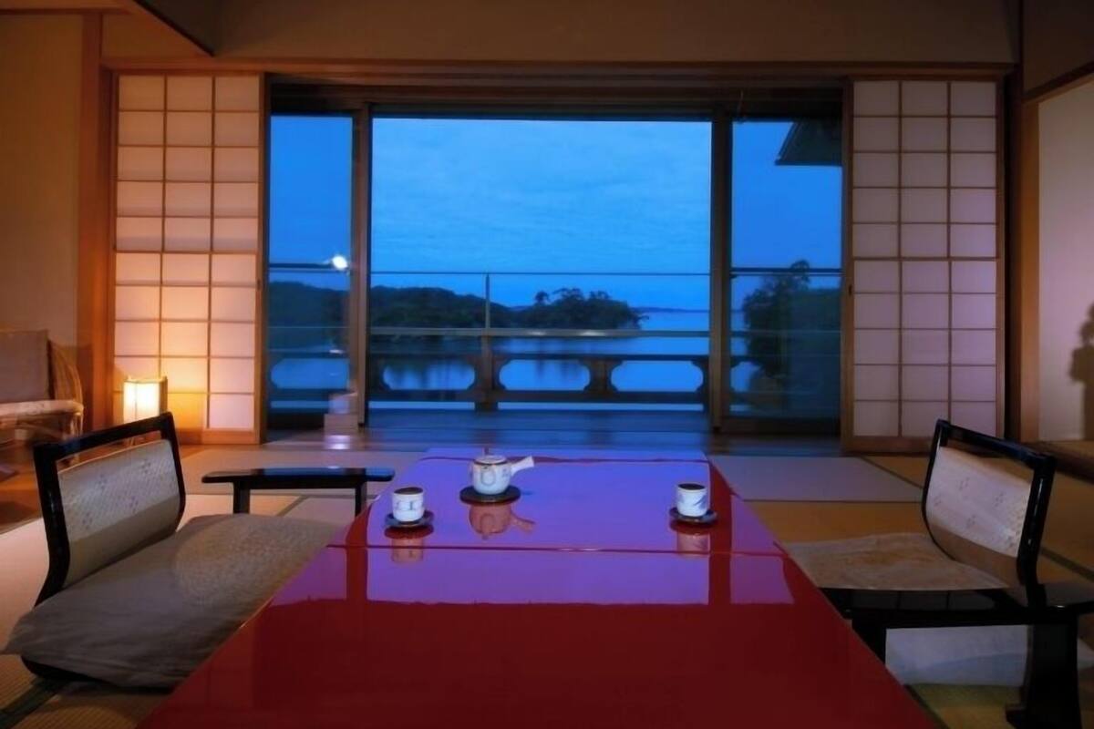 Una habitación con un gran ventanal corredero, una mesa roja con dos tazas y una tetera, y puertas correderas tradicionales japonesas.