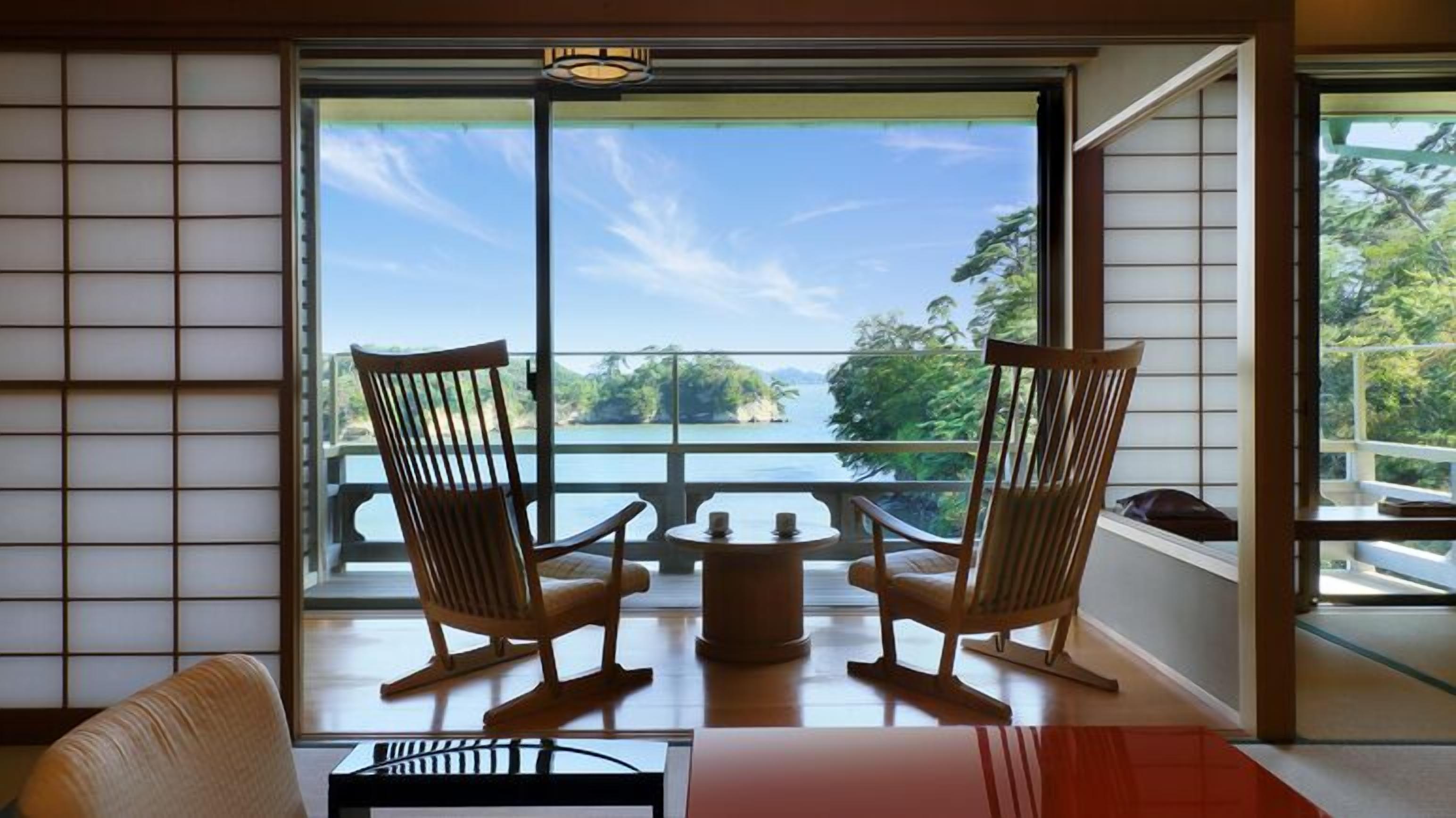 Una habitación con dos mecedoras de madera orientadas a un balcón con vistas a un lago y árboles.