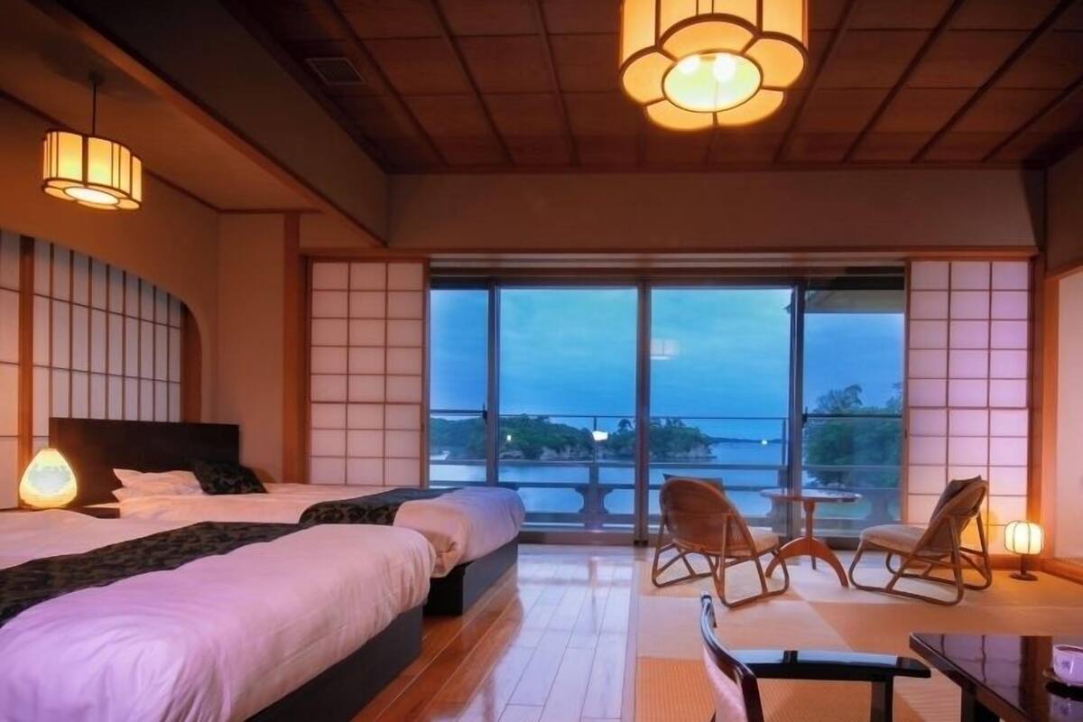 Habitación de hotel con una cama grande, zona de estar con sillas, vista al mar y puertas correderas tradicionales japonesas.