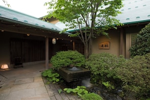 A traditional Japanese building with a stone pathway leading to a wooden gate, surrounded by lush greenery and a small stone fountain. - Matsushima Sakan Shoan (Matsushima)