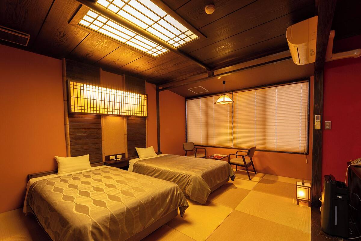 A hotel room with two beds, a small table, chairs, and a window with blinds.