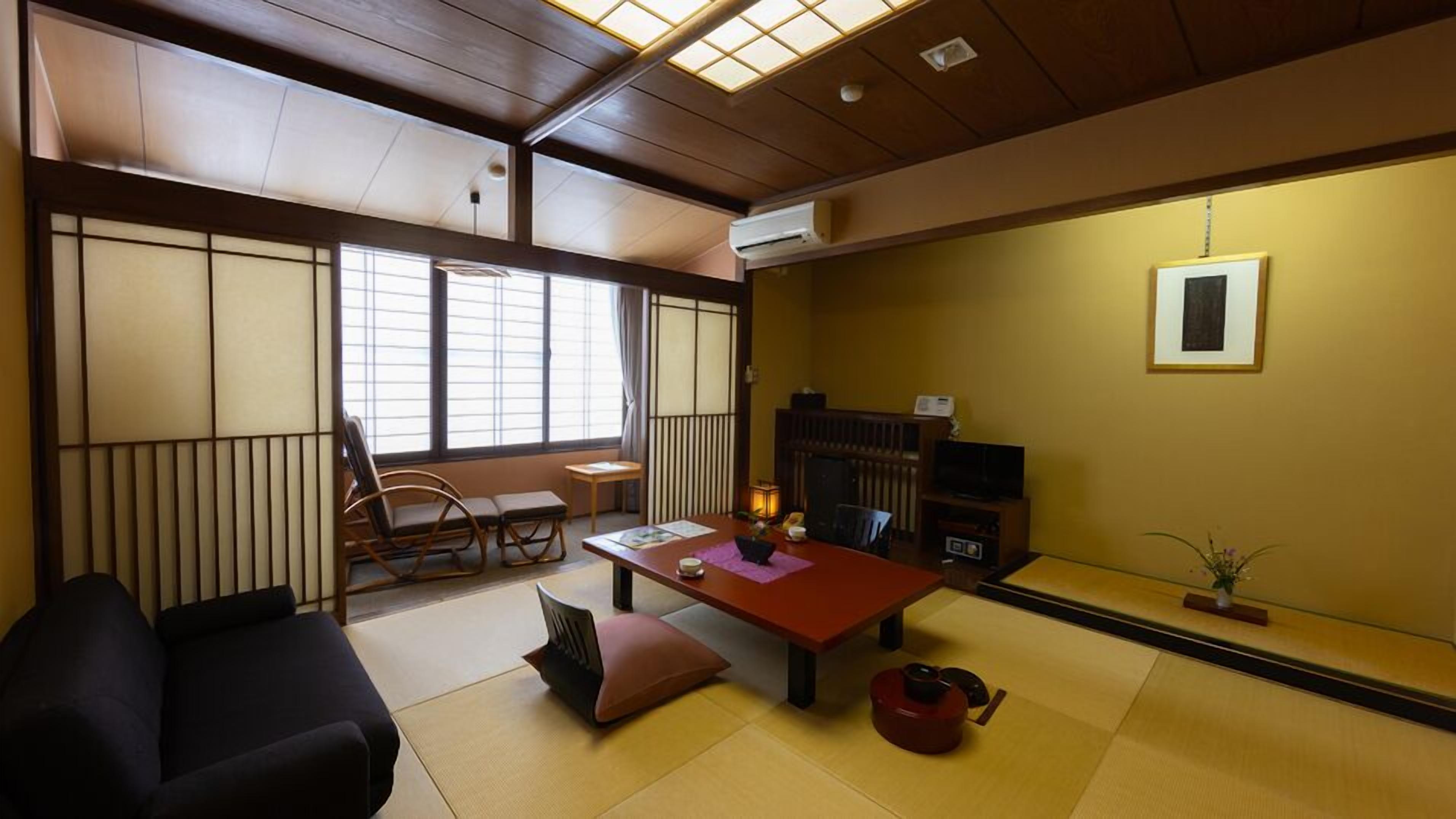 Une pièce de style japonais traditionnel, avec un sol recouvert de tatamis, une table basse entourée de coussins, un canapé noir, un téléviseur et une cheminée.