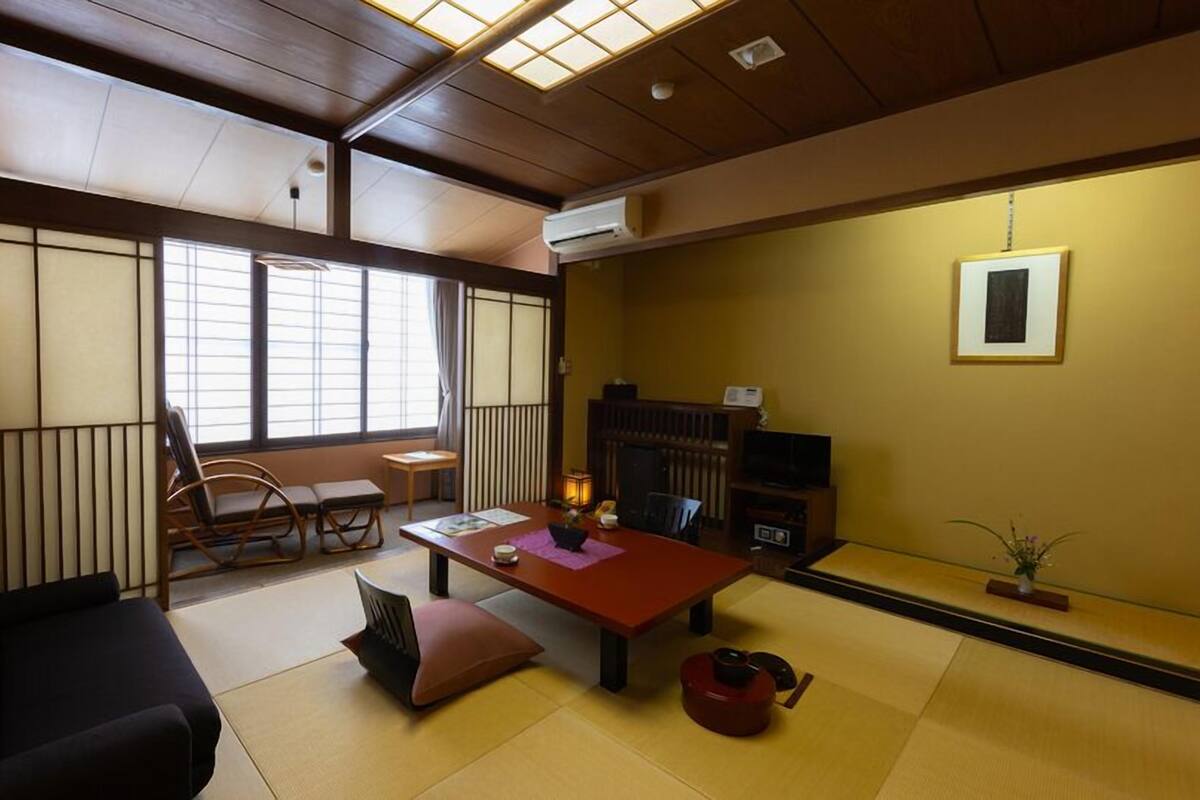 A traditional Japanese room with tatami flooring, a low table with cushions, a black sofa, a television, and a fireplace.