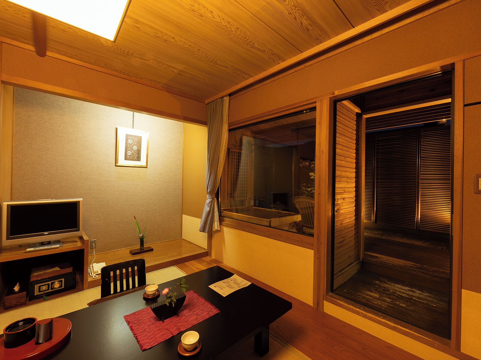 A traditional Japanese-style room with tatami flooring, a low table with a red mat, a TV, and a sliding door leading to a garden.