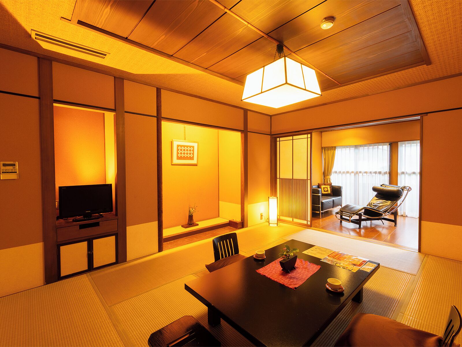 Una habitación de estilo japonés tradicional con suelo de tatami, una mesa baja con un posavasos, un televisor y una puerta corrediza que da a un balcón con un sofá y una silla.